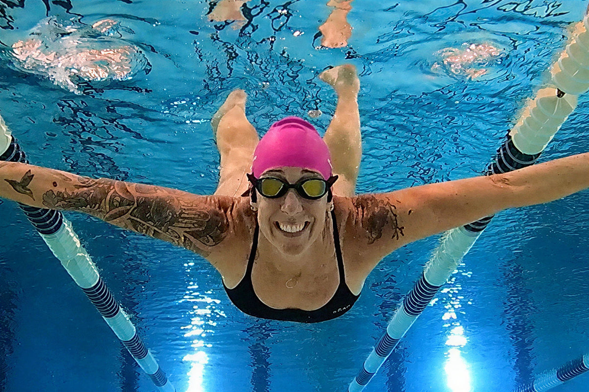 A smiling woman underwater wearing black tinted Basilisk goggles with her arms spread wide 