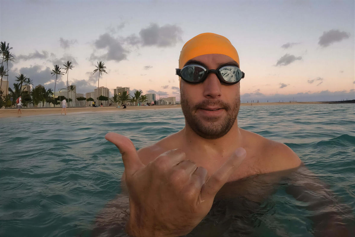 A man wearing S&P goggles and an orange swim cap makes a hang-loose sign with his hand while floating in the ocean off Waikiki Beach